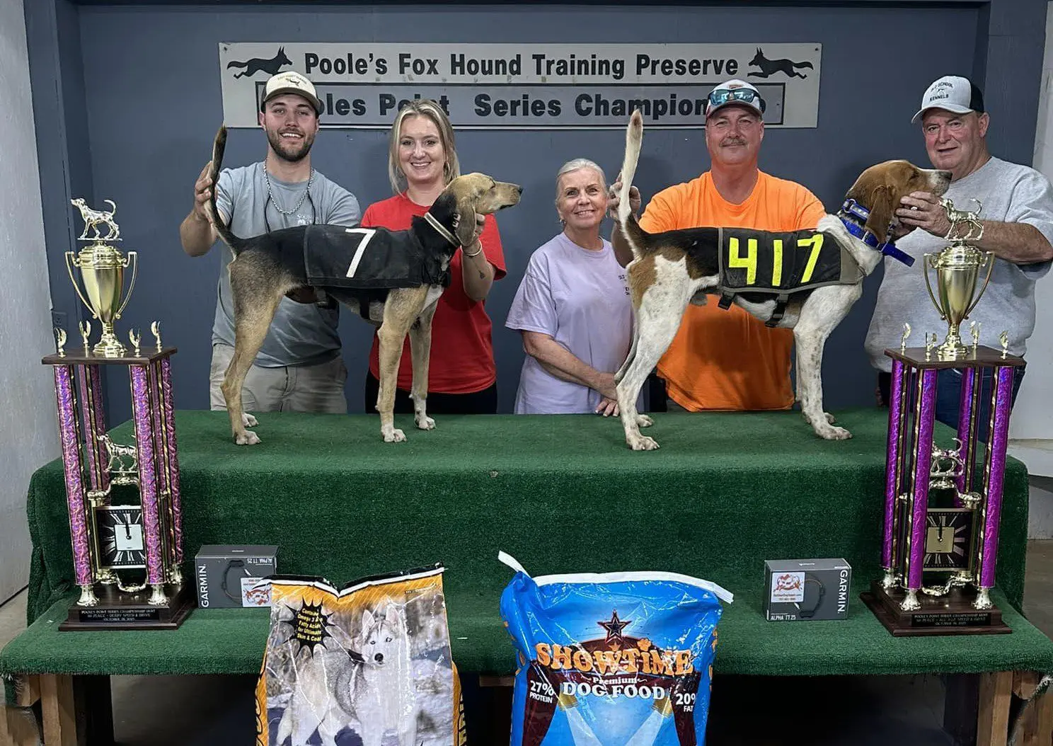 Two dogs with their handlers at a dog show podium with prizes.