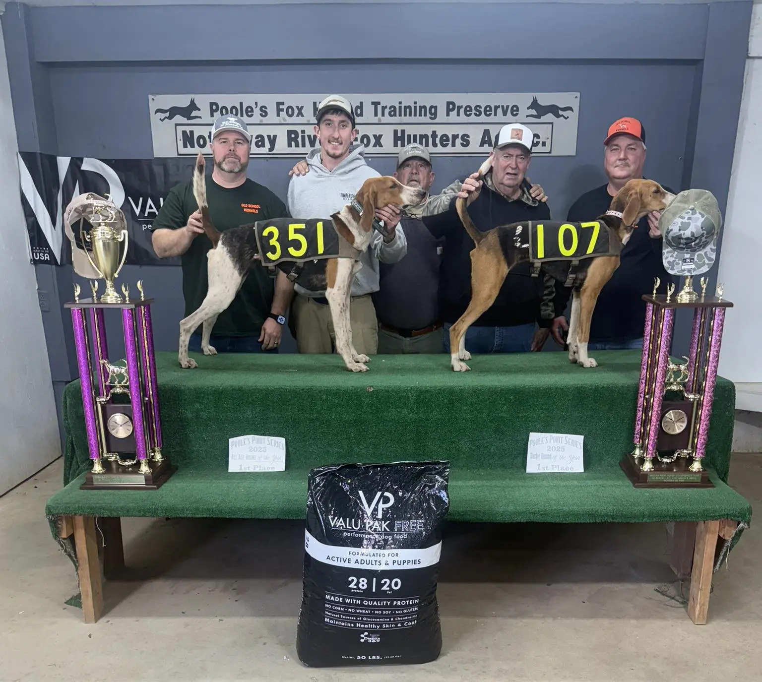 Three dogs wearing numbered vests on a bench with trophies and a bag of dog food.