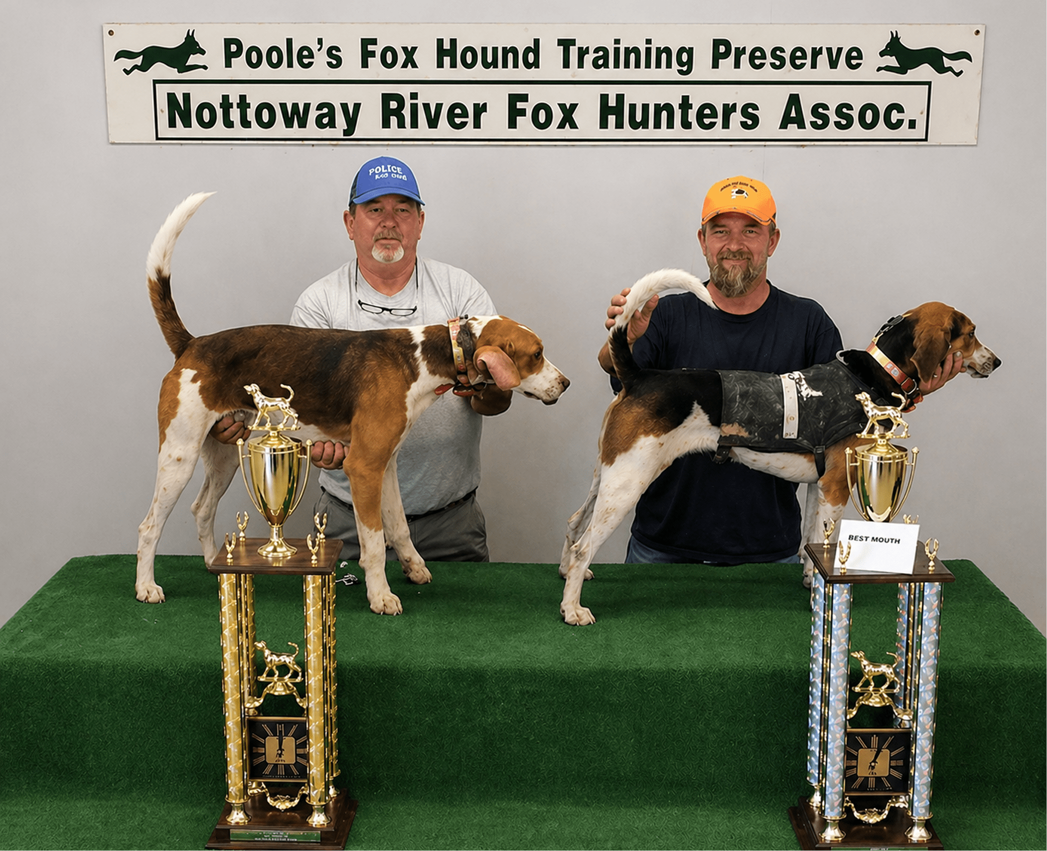 Two men proudly pose with their Fox Hounds and trophies at a hunting association event.
