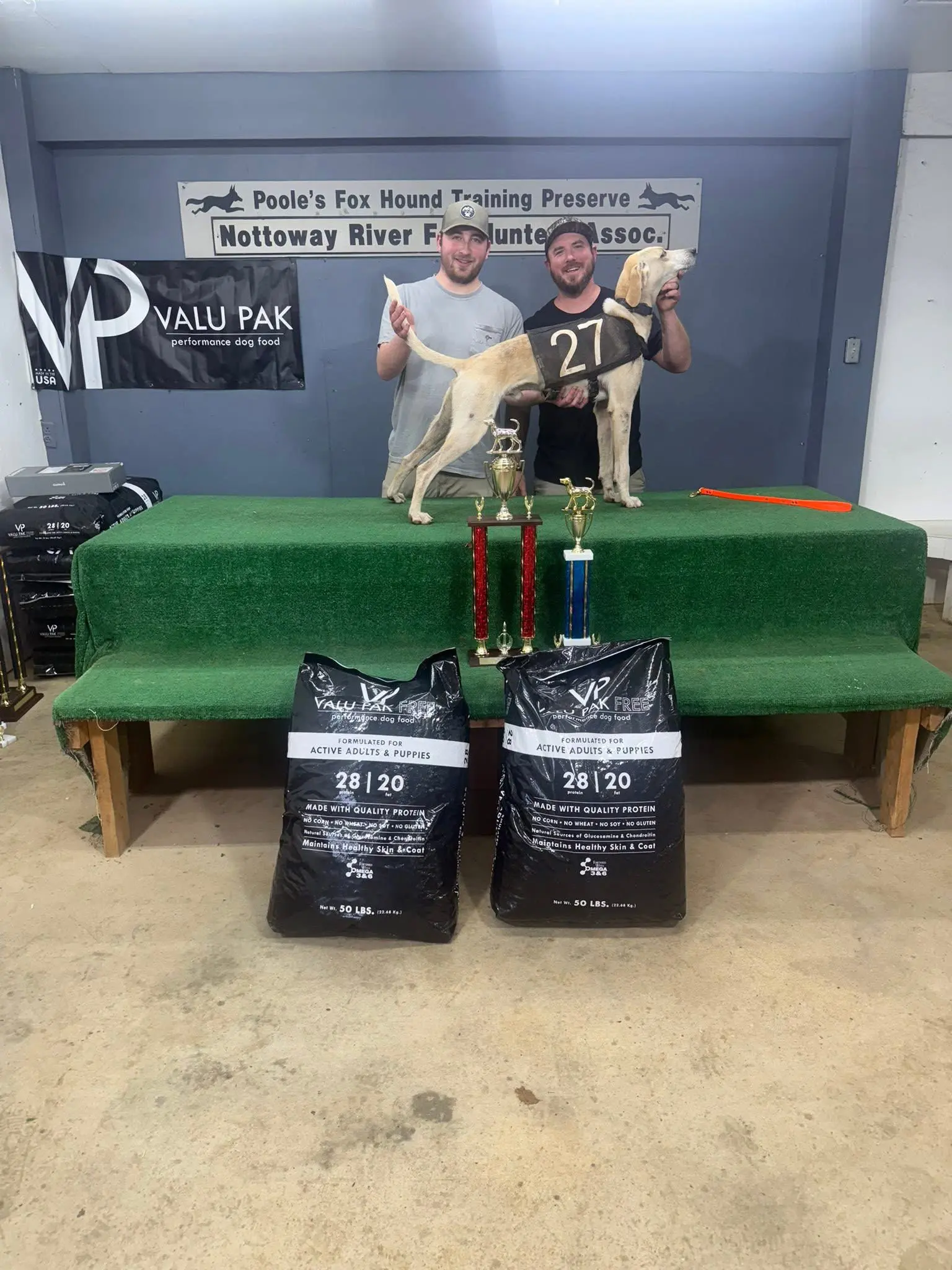 Two men with dog and trophies on table.