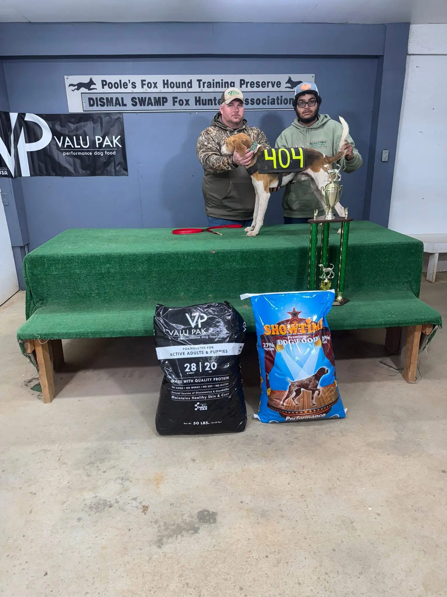 Two men with a dog and trophies.