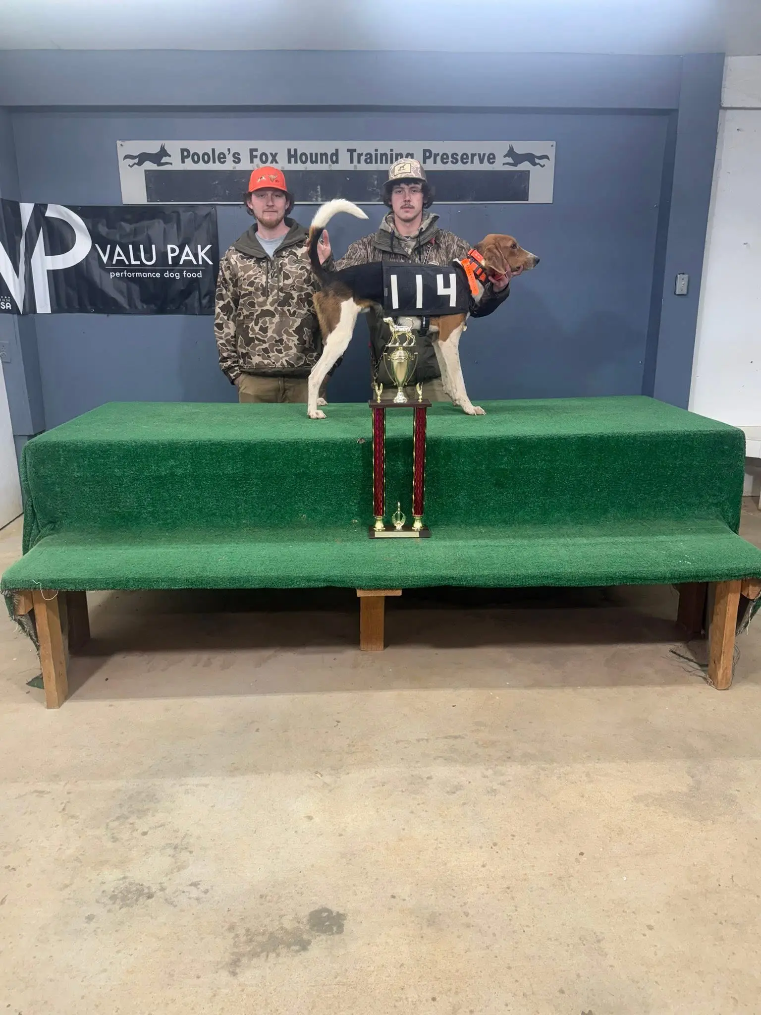 Two men with dog and trophy on table.